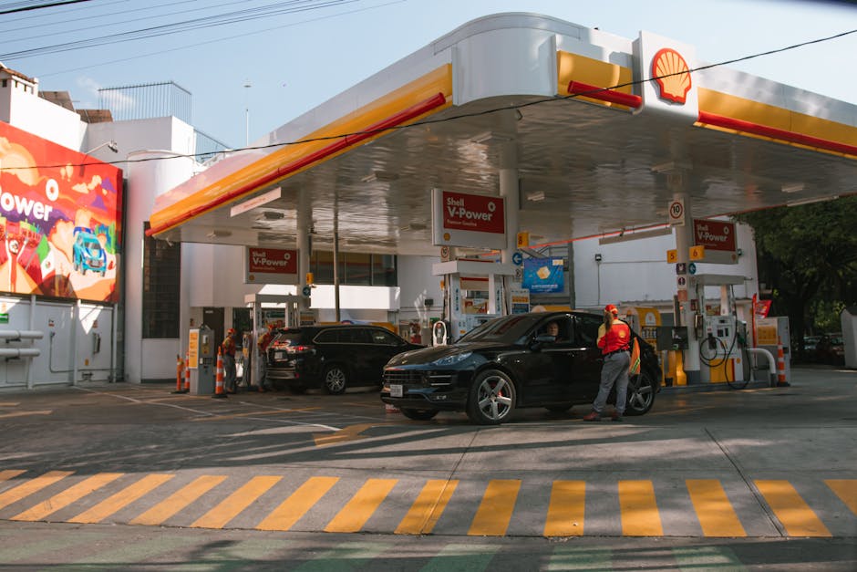 A Shell gas station with vehicles refueling and attendants assisting drivers on a sunny day.